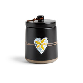 A round black ceramic poured candle with a tan textured base, a lid and a white raised heart on the front with a yellow floral illustration.