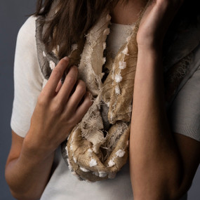 A tan infinity scarf with white strings running through it and a short fringed edge, being worn by a woman in a white short sleeve sweater.