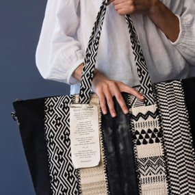 A woman holding a black and white patterned large tote bag by the handles in front of a blue gray background.