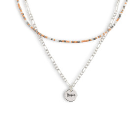 A layered necklace made up of a silver chain with a round silver charm that says "Brave" and a layer of orange, gray and white small beads.