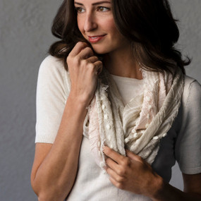 A blush colored textured infinity scarf with white threads running through it and a short fringed edge, being worn by a woman in a short sleeve white sweater.