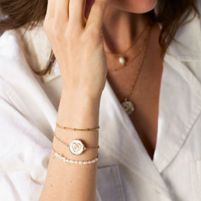 Close view of a woman wearing a multilayered gold chain and white bead bracelet with a round heart charm.