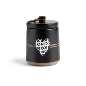 A round black ceramic poured candle with a tan textured base, a lid and a white raised heart on the front with a black X and O illustration.