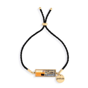 An adjustable black string bracelet with a horizontal gold charm with orange and brown abstract artwork inspired by ArtLifting artist Cheryl Kinderknecht. The bracelet also has a small round gold token that says "gratitude".