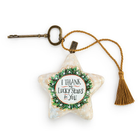 A cream star shaped sculpture with the saying "I Thank my Lucky Stars for You" inside an illustrated wreath. The star has a gold tassel and gold key attached.
