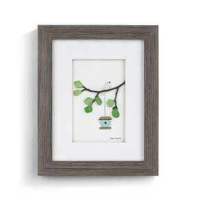 A gray wood framed image of a birdhouse hanging from a tree branch with a white bird perched on it made of pebbles.