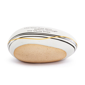 A white and tan stoneware rock with black and gold horizontal lines and "The love they gave echoes through our whole lives" on the top.