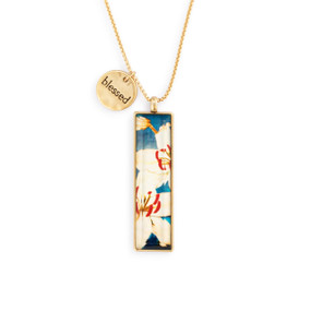 An adjustable gold chain necklace with a vertical gold charm with a white floral pattern artwork inspired by ArtLifting artist Midori. The necklace also has a small round gold token that says "blessed".