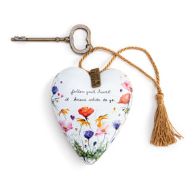 A white heart shaped sculpture with the image of wildflowers that says "follow your heart it knows where to go." The heart has a a key and gold tassel attached.