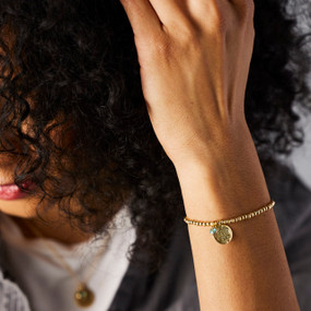 Close up view showing a woman wearing a gold chain bracelet with a round flower charm and stone representing the month of March.