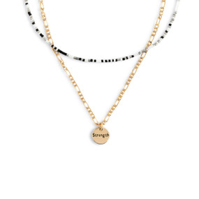 A layered necklace made of a gold chain with a round gold charm that says "Strength" and a layer of black, gray and white small beads.