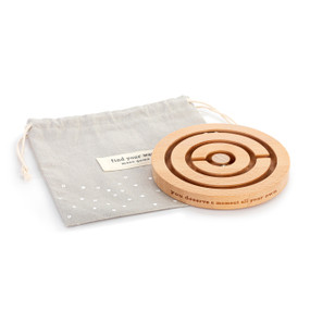 A light wood round maze game with small round metal beads to move around the maze. The side of the maze says "you deserve a moment all your own", displayed next to its light gray fabric storage bag.