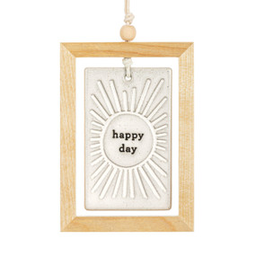 A hanging cream ceramic plaque with a sun and the saying "happy day" suspended inside a light wood open frame.