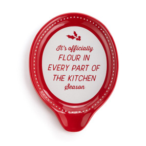 Top down view of a red ceramic spoon rest with white dots and the saying "It's officially Flour In Every Part of the Kitchen Season".