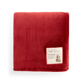 A large red blanket folded and has a fabric tag that says "What should we do today?" asked Piglet. "Nothing," said Pooh. "And let's do it all day." with an image of Piglet and Pooh.