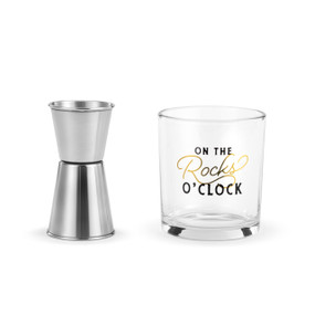 A clear rocks glass with the saying "On The Rocks O'Clock" next to a silver metal jigger.