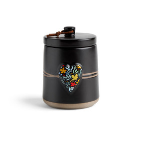 A round black ceramic poured candle with a tan textured base, a lid and a black raised heart on the front with a colorful floral illustration.