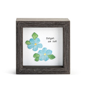 A gray wood shadow box with a pebble art inside of two blue flowers that says "forget me not".