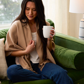 A woman in a camel colored wrap sitting on a green couch holding a mug.