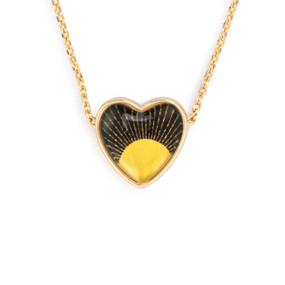 Detail view of the charm on an adjustable gold chain necklace with a heart shaped charm of a yellow sun on a dark background.