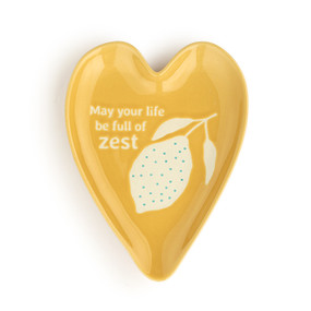 A yellow heart shaped ceramic spoon rest with a lemon illustration and the saying "May your life be full of zest" on it.