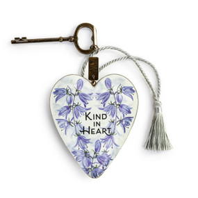 A heart shaped white sculpture with illustrated purple flowers with a silver tassel and metal key attached. The heart has the saying "Kind in Heart".