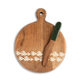 A round wood serving board with a small handle at the top and white lines like pine needles at the bottom. A small wood spreader with a dark green plastic handle is laid on top.