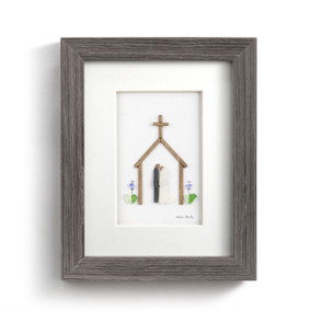 A wall art in a gray wood frame. The image is pebbles and wood arranged to look like a couple inside a wedding chapel.