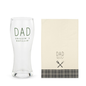 A clear pilsner glass that says "Dad Grillin' & Refillin'" next to a cream towel with the same saying and a plaid pattern at the bottom.