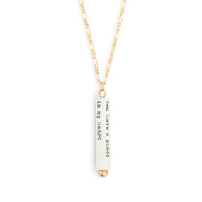 A gold chain necklace with a white scroll charm. The scroll has a saying from F. Scott Fitzgerald.