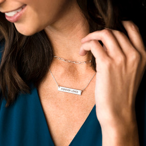 Close up of a woman wearing a silver chain necklace with a bar charm that says "Forever Loved".