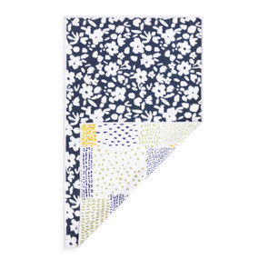 A rectangular double sided towel. One side is dark blue with illustrated white flowers and the other is white with a patchwork pattern, displayed with the patchwork corner turned back.
