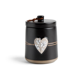 A round black ceramic poured candle with a tan textured base, a lid and a light brown raised heart on the front with a white floral illustration.