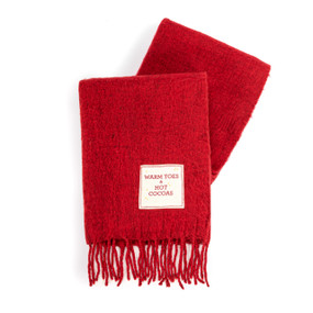 A red fringed blanket with a cream fabric patch that says "Warm Toes & Hot Cocoas".