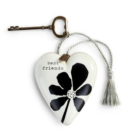 A heart shaped sculpture that is white and says "best friends" in black with a black flower. The sculpture has a gold key and silver tassel attached.