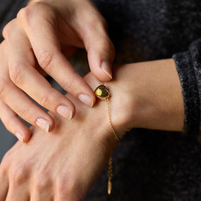 Close up of a woman wearing a gold chain bracelet with a heart shaped charm with an illustration of the sun.