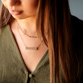 Close up of a woman wearing a gold necklace with two different chains and a gold bar that says "Forever Loved".