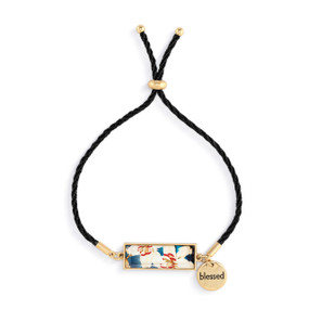 An adjustable black string bracelet with a horizontal gold charm with white floral artwork inspired by ArtLifting artist Midori. The bracelet also has a small round gold token that says "blessed".