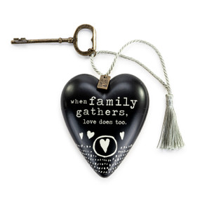 A heart shaped sculpture that is black and says "when family gathers, love does too." in white with white hearts. The sculpture has a gold key and silver tassel attached.