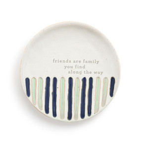 A small white round ceramic trinket dish with vertical green and blue stripes on the bottom with the saying "friends are family you find along the way" in the middle.