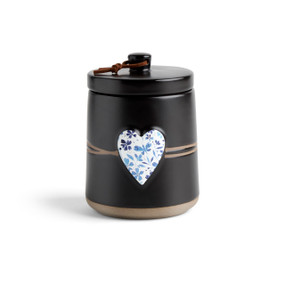 A round black ceramic poured candle with a tan textured base, a lid and a white raised heart on the front with a blue floral illustration.