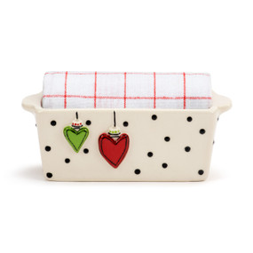 A mini cream ceramic loaf pan with black dots and a raised red and green heart ornament on the front and white striped towel rolled inside.