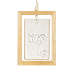 A hanging cream ceramic plaque with XOXO and a heart suspended inside a light wood open frame.