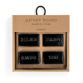 A set of four black ceramic square napkin rings. Each ring has a different word on it "Delish, Enjoy, Savor and Yum", displayed in a packaging box.