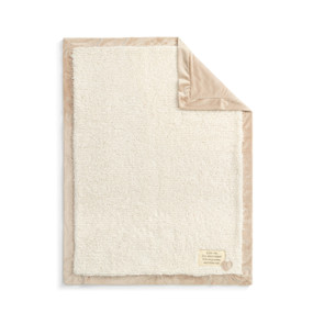 A plush cream blanket with a tan edge and back. There is a fabric patch in the bottom right corner.