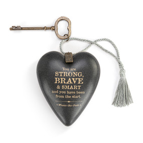 Heart shaped sculpture with a silver tassel and metal key attached. The heart has a dark gray background and says "You are Strong, Brave & Smart and you have been from the start".
