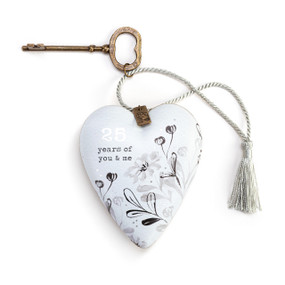 A white heart shaped sculpture that says "25 years of you & me" with gray artistic flowers. The heart has a silver tassel and gold key attached.