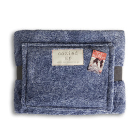 A blue lap blanket with a hand pocket. The fabric patch says "cozied up with no place to go", wrapped in a gray ribbon with a product tag attached.