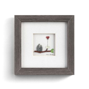 A square wall art in a gray wood frame. The image is pebbles arranged to look like a couple sitting together with a red heart balloon.