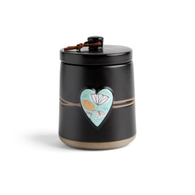 A round black ceramic poured candle with a tan textured base, a lid and a light green raised heart on the front with a floral illustration.